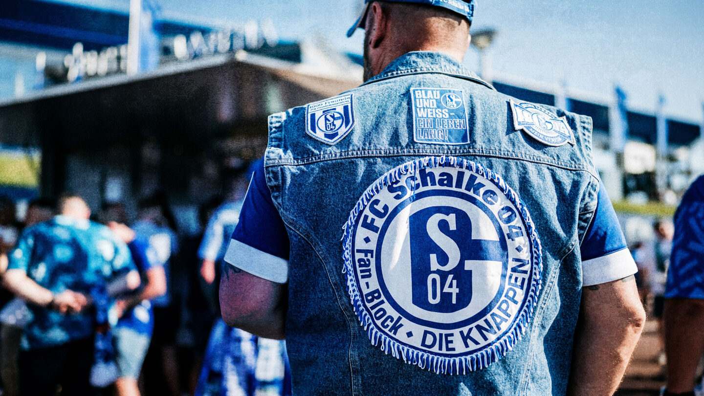 Schalkefan in einer Jeanskutte mit großem FC Schalke 04-Logo auf dem Rücken, steht vor dem Stadion zusammen mit anderen Fans.