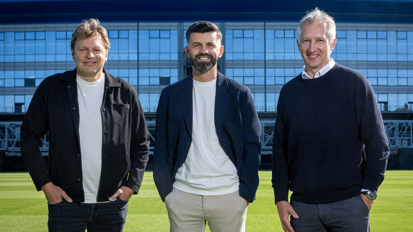 Youri Mulder, Miron Muslic und Frank Baumann stehen gemeinsam auf dem herausgefahrenen Rasen des Stadions. Im Hintergrund ist die VELTINS-Arena zu sehen.