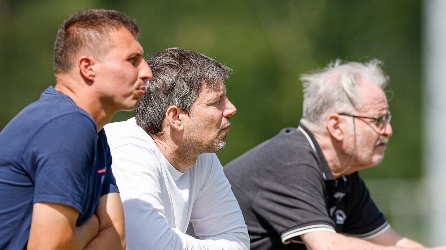 Drei Männer, darunter Raffael Tonello, lehnen an der Bande und verfolgen ein Fußballspiel.
