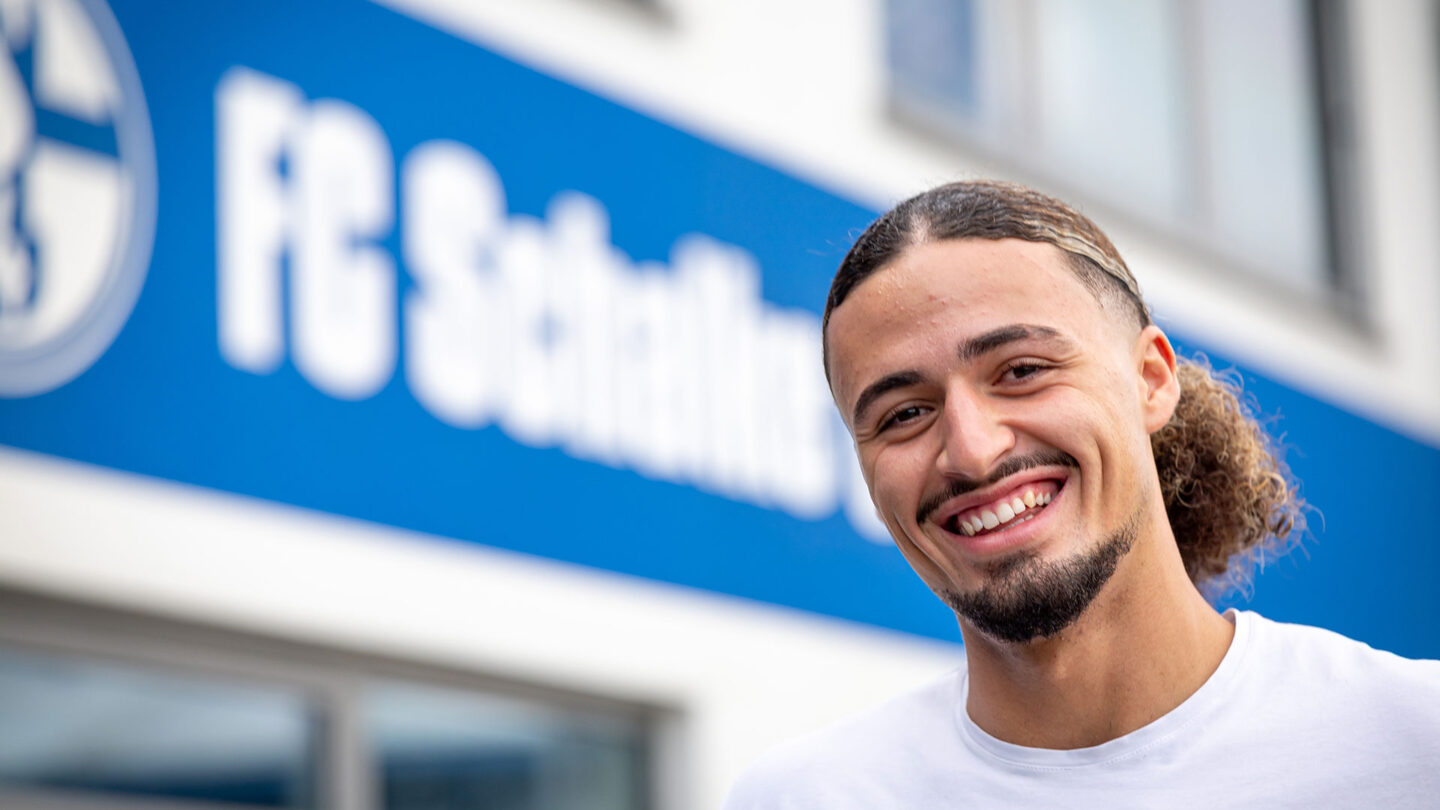 Taylan Bult steht lachend vor dem Profileistungszentrum. Im Hintergrund ist der Schriftzug FC Schalke 04 zu sehen.