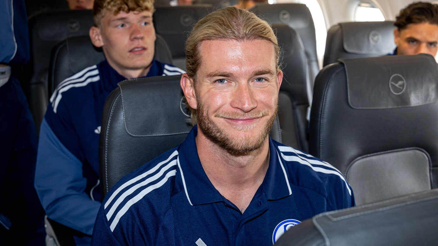Loris Karius sitzt im Flugzeug und schaut in die Kamera. Im Hintergrund ist Johannes Siebeking zu sehen.