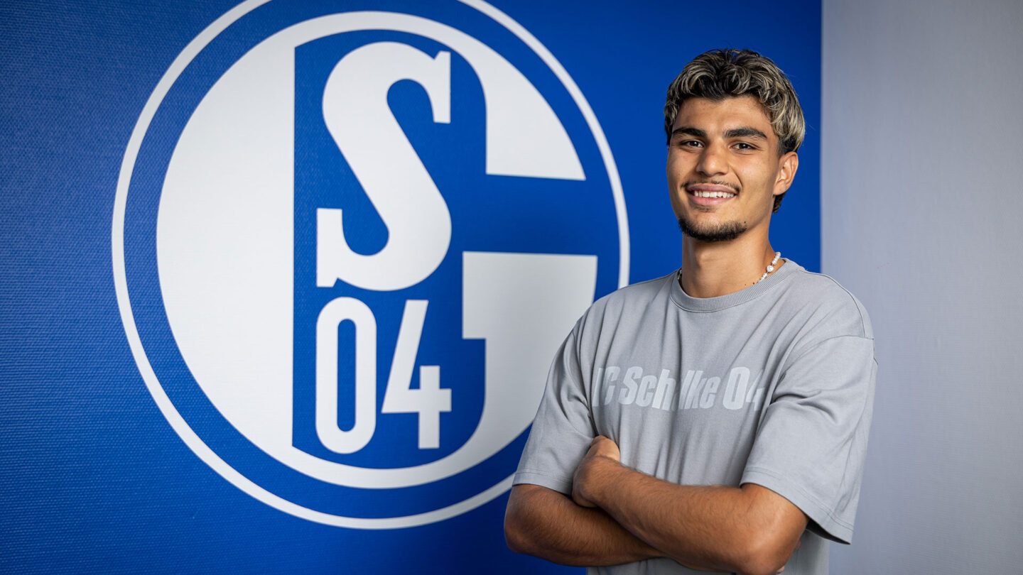 Mertcan Ayhan steht mit verschränkten Armen vor einem großen S04-Logo an der Wand.