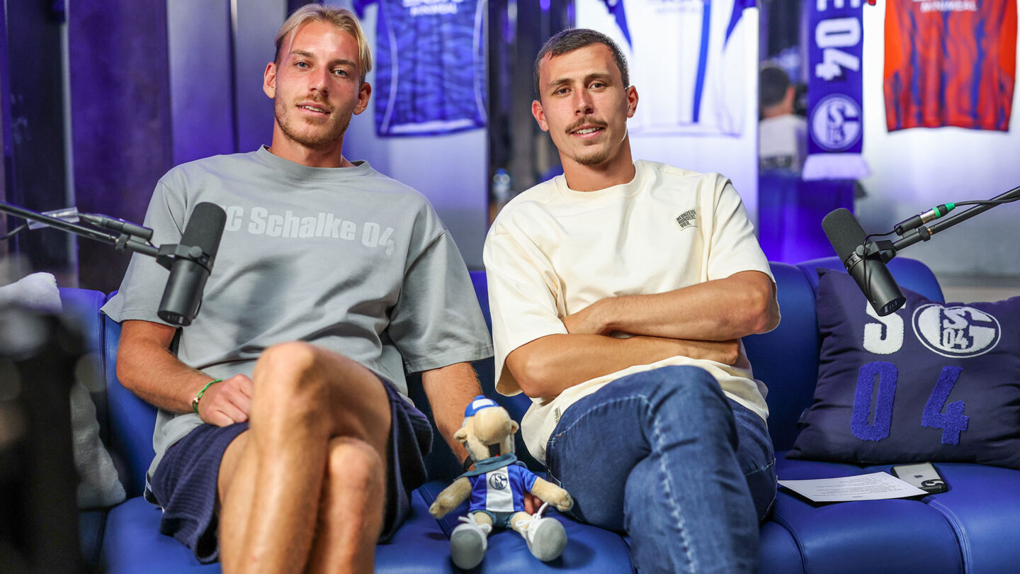 Timo Becker und Ron Schallenberg sitzen in der Kabine. Hinter ihnen sind Trikots zu sehen, vor ihnen ist jeweils ein Mikrofon zu sehen.