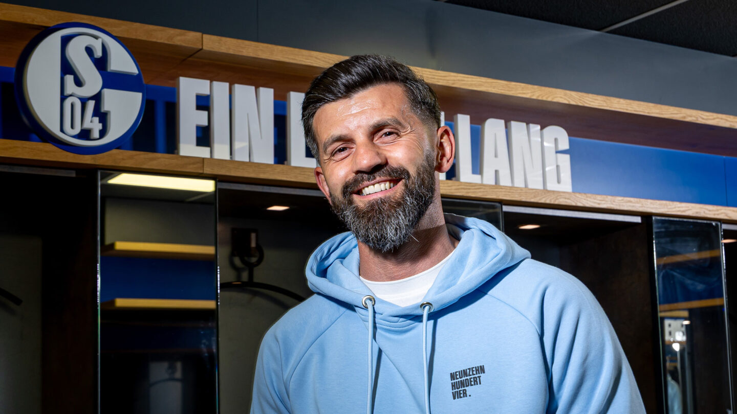 Miron Muslic steht lachend in der Umkleidekabine in der VELTINS-Arena. Hinter ihm befinden sich mehrere Spinde sowie das S04-Logo mit dem Schriftzug ein Leben lang.
